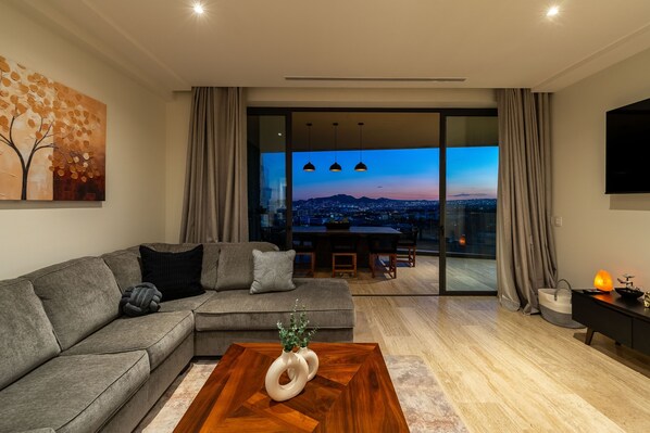 Ein modernes Wohnzimmer mit Sofa, Couchtisch, einem Gemälde, einem Essbereich und Blick auf die Stadt im Abendlicht.