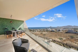 Ein Balkon mit Blick auf das Meer und ferne Berge, eine Essecke mit Stühlen und Tisch sowie eine Lounge mit einer Couch.