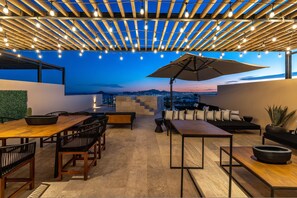 En udendørs terrasse med et træpergola, lyskæder, et spiseområde med stole og borde samt en lounge med sofaer og en stor parasol.