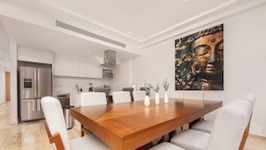 A modern dining area with a wooden table and white upholstered chairs, a kitchen in the background, and a large Buddha painting on the wall. - Mistiq Views in Cabo San Lucas (Cabo San Lucas)