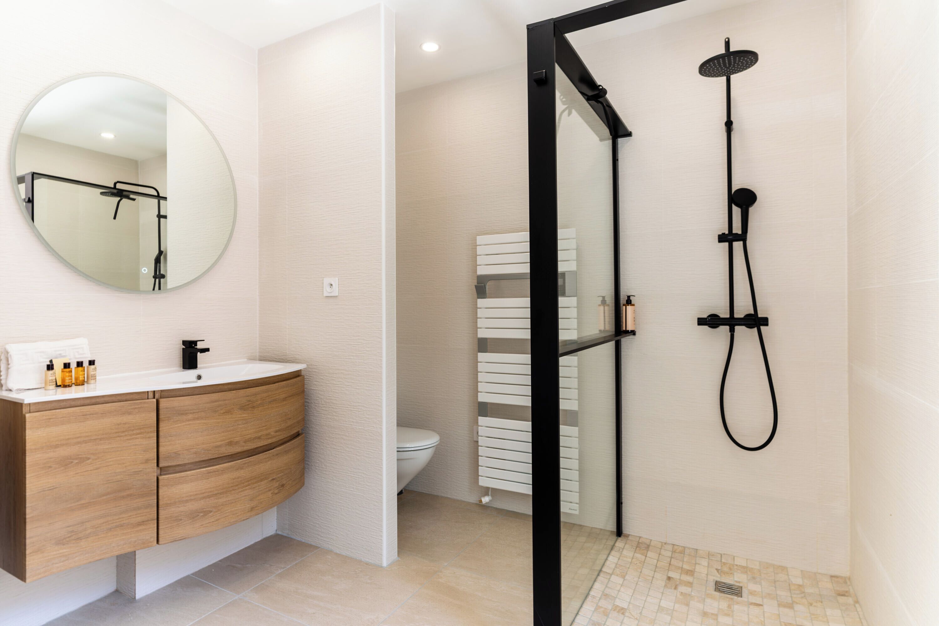 A bathroom with a wooden vanity, a round mirror, a toilet, and a shower area.