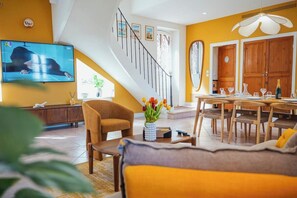 En stue med sofa, sofabord, fjernsyn, trappe og spiseområde med bord og stole.