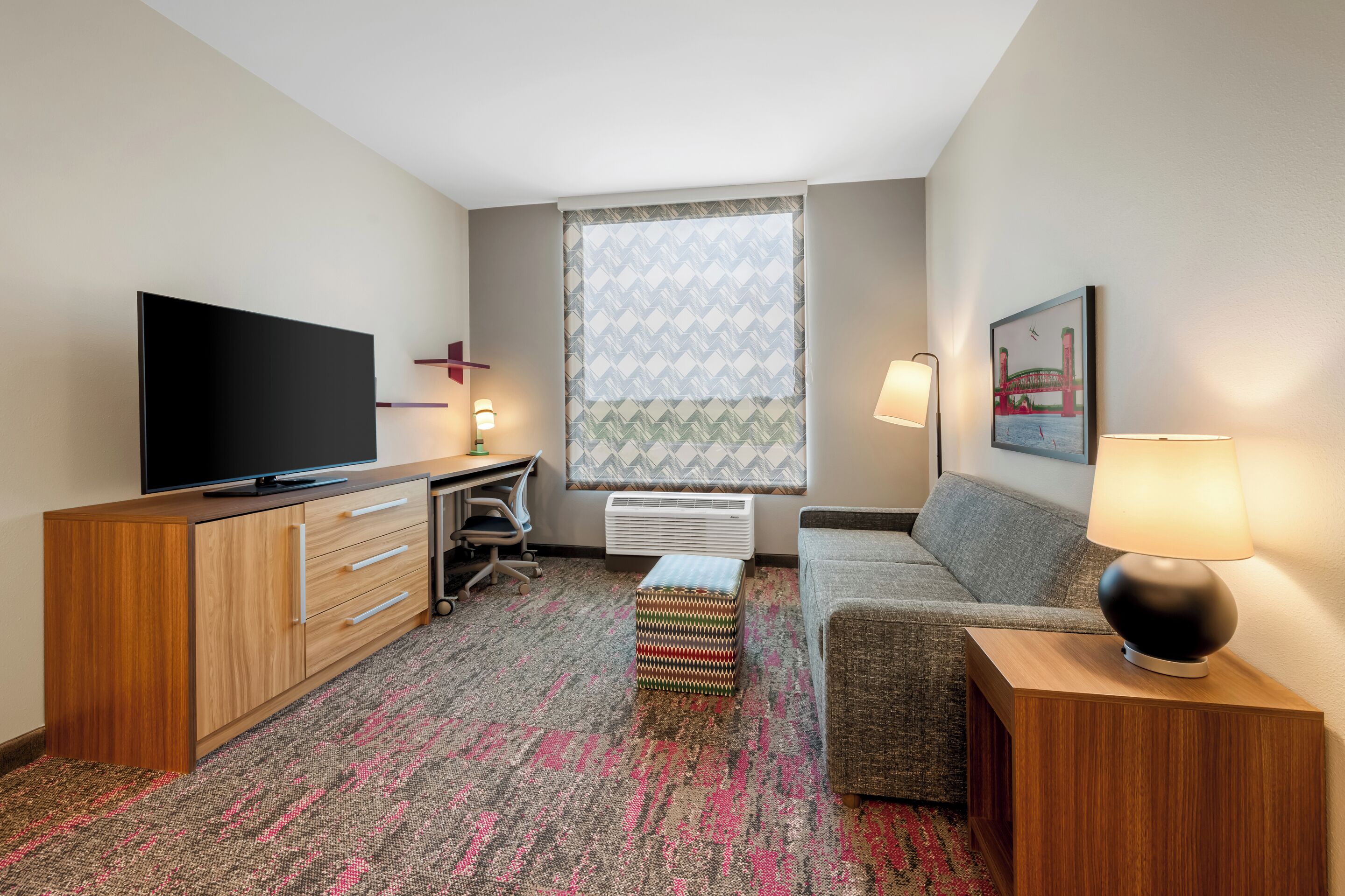 A hotel room with a flat-screen TV, a desk with a chair, a sofa, a patterned rug, and a window with patterned curtains.