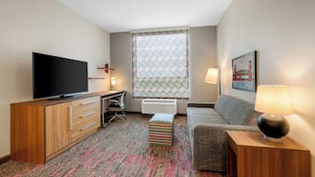 A hotel room with a flat-screen TV, a desk with a chair, a sofa, a patterned rug, and a window with patterned curtains.