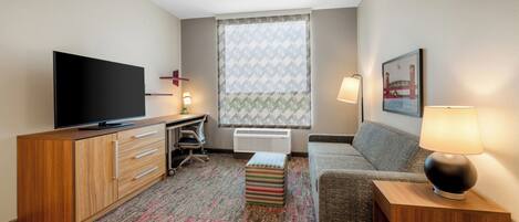 A hotel room with a flat-screen TV, a desk with a chair, a sofa, a patterned rug, and a window with patterned curtains.