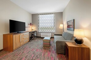 A hotel room with a flat-screen TV, a desk with a chair, a sofa, a patterned rug, and a window with patterned curtains.