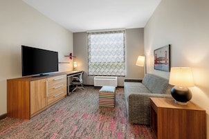 Habitación de hotel con televisión de pantalla plana, escritorio con silla, sofá, alfombra estampada y ventana con cortinas decoradas.