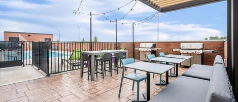 Un'area patio esterna con tavoli e sedie, un barbecue e una piscina sullo sfondo.