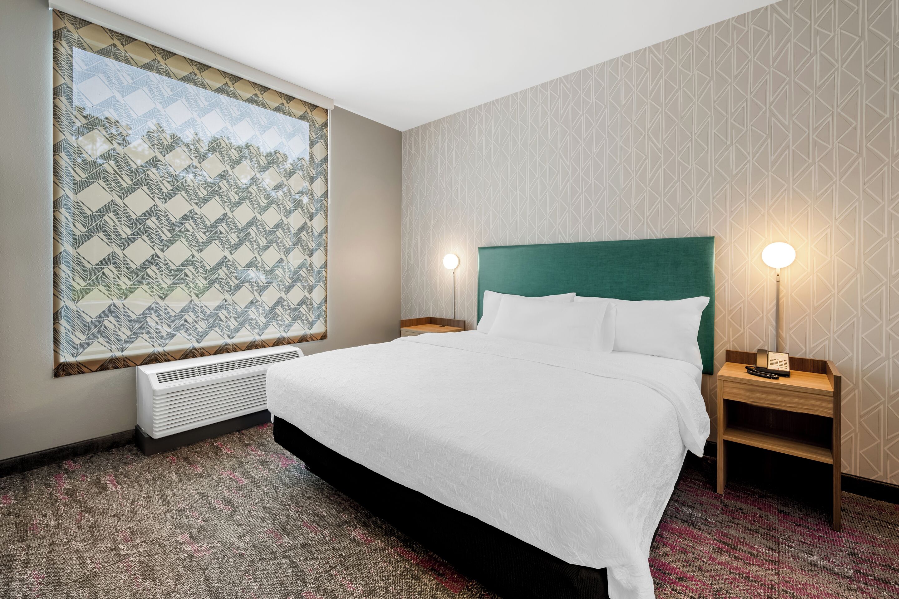 A hotel room with a large bed, two bedside tables with lamps, a large window with a geometric view, and a wall-mounted air conditioner.