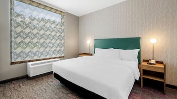 A hotel room with a large bed, two bedside tables with lamps, a large window with a geometric view, and a wall-mounted air conditioner.