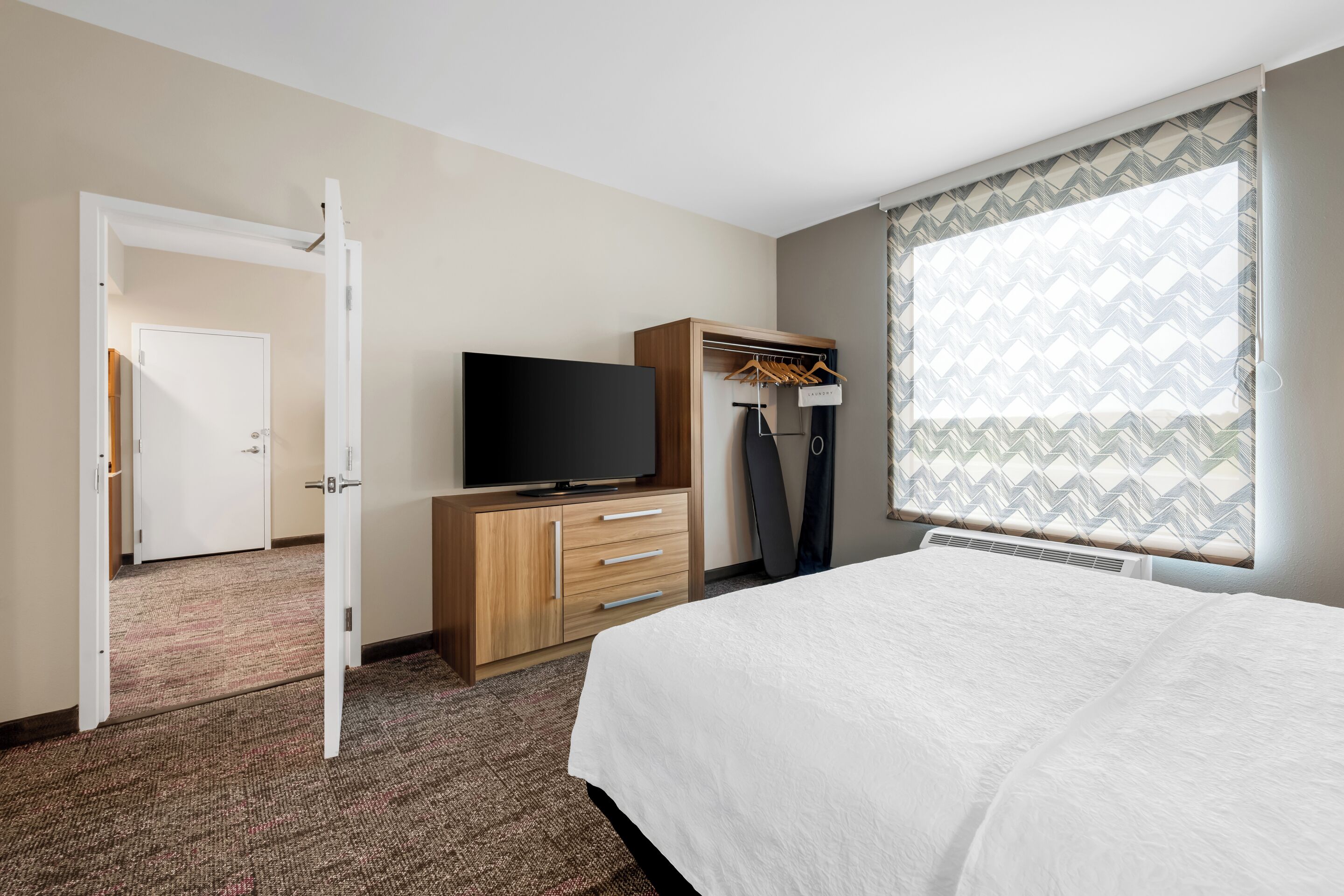 Una habitación de hotel con cama, televisor, armario y ventana con cortinas estampadas.