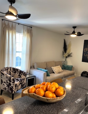 Ein modernes Wohnzimmer mit Sofa, einem gemusterten Sessel, einer Holzschale mit Orangen auf einer Granitarbeitsplatte und Deckenventilatoren.