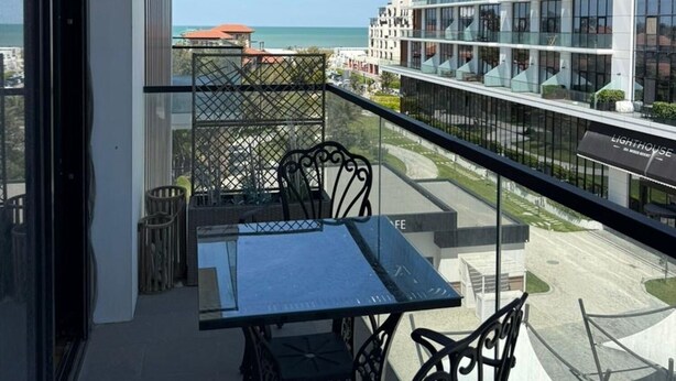 Varanda com mesa e cadeiras, vista para um prédio moderno e o mar.
