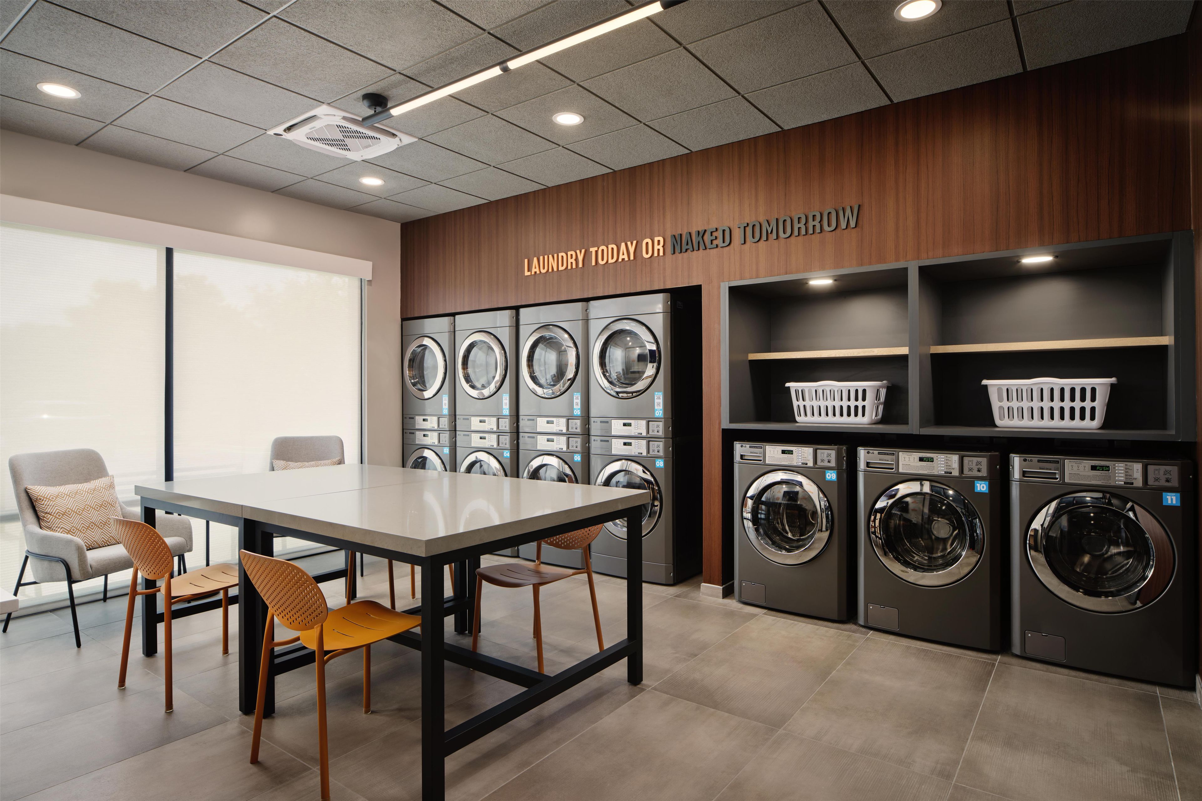 A laundry facility with multiple washing machines, a table with chairs, and a sign above the machines.