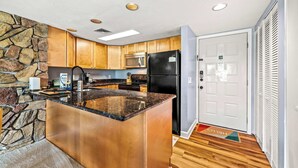 A modern kitchen with granite countertops, wooden cabinets, a stone accent wall, a black refrigerator, and a microwave. - Mountain View Condo w/ Indoor Pool & Golf Access (Gatlinburg)