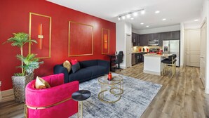 Ein modernes Wohnzimmer mit einer roten Akzentwand, einem schwarzen Sofa, einem pinken Sessel, einem runden Couchtisch und einer Küche im Hintergrund.