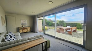 A modern living room with a sofa, a wooden coffee table, a cabinet, and a large sliding glass door leading to an outdoor patio with a table and chairs. - Spacious 4 bedroom holiday home (Lake Tekapo)