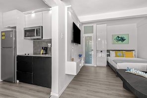 Una habitación de hotel moderna con kitchenette, un televisor de pantalla plana, una cama con un cartel de 'Welcome' y un cuadro en la pared.