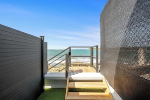 Una escalera de madera que baja a una playa, con baranda de metal, una pared gris y una malla protectora.