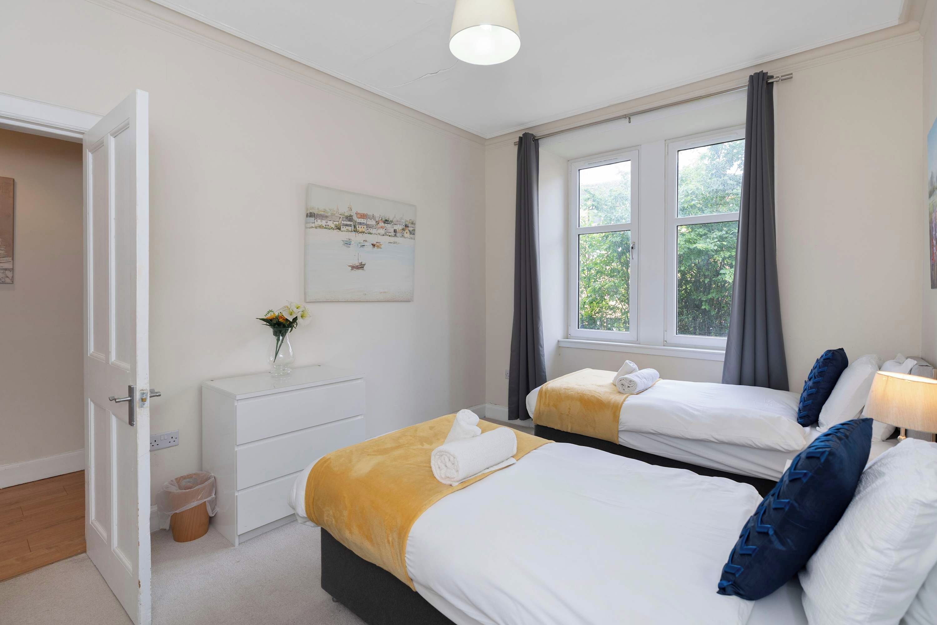 A hotel room with two beds, a white dresser, a painting, and a window with curtains.