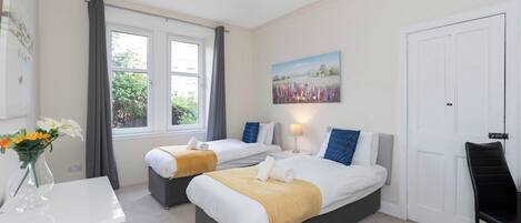 A hotel room with two beds, a desk, a chair, a painting on the wall, and a window with curtains.
