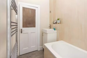 A bathroom with a white bathtub, a white sink, a door with a frosted glass panel, and a small shelf with a plant and toiletries.