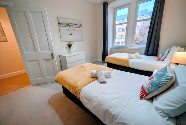 A hotel room with two beds, a nightstand, a window with curtains, and a wall-mounted artwork.