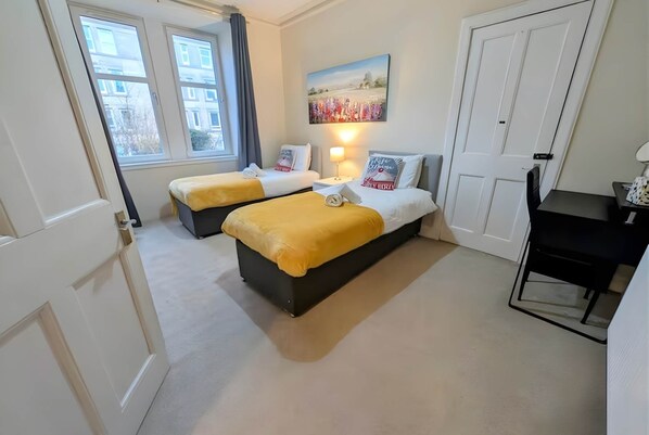 A hotel room with two beds, a desk, a chair, a painting on the wall, and a window with curtains.