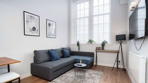 Een moderne woonkamer met een grijze hoekbank, een klein rond salontafeltje, een staande vloerlamp en een groot raam met zonwering.