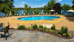 A circular pool with a yellow edge, surrounded by lounge chairs and umbrellas, with a view of a lake and trees in the background. - Four-Season Lake Winnipesaukee Retreat (Laconia)