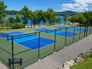 Een tennisbaan met een net, omringd door een hek, en een meer op de achtergrond.