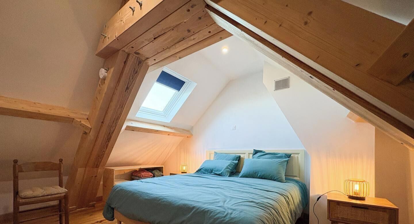 Un dormitorio con techo inclinado, un tragaluz, una cama con ropa de cama azul, una silla y una mesita de noche con una lámpara.