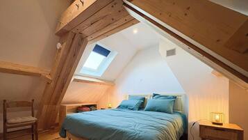 Un dormitorio con techo inclinado, un tragaluz, una cama con ropa de cama azul, una silla y una mesita de noche con una lámpara.