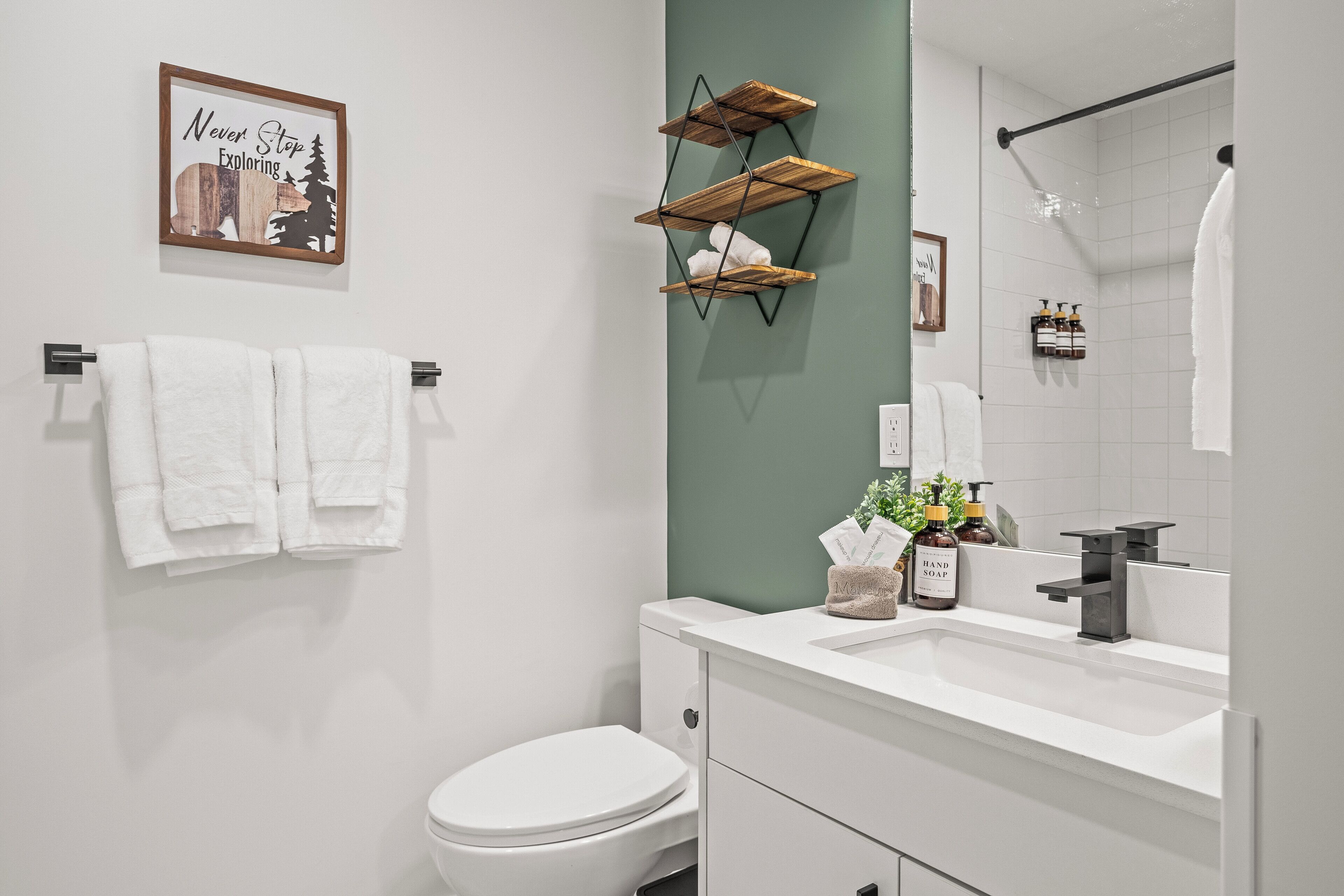 A bathroom with a white toilet, a white sink with a modern faucet, and a shower area. There is a framed picture on the wall and a towel rack with two towels.