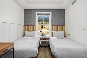 A hotel room with two beds, a window with a view of mountains and trees, a wooden nightstand with a lamp, and a white wardrobe.
