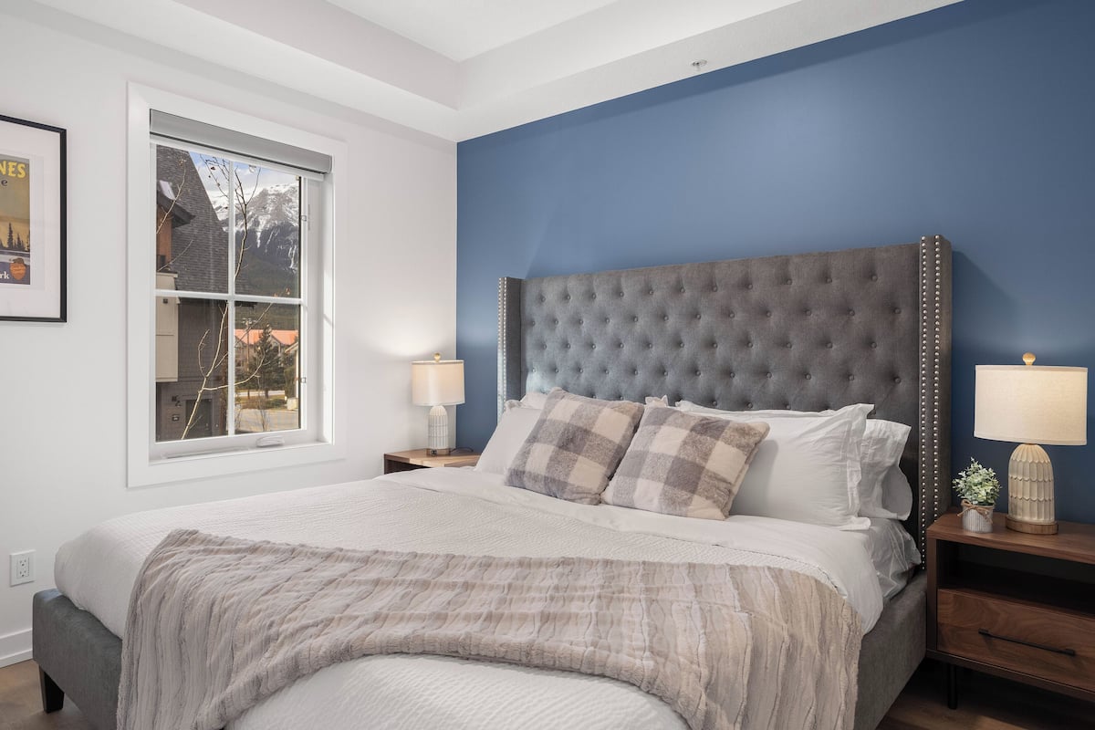 A bedroom with a large bed, a window with a mountain view, a blue accent wall, and a tufted headboard.
