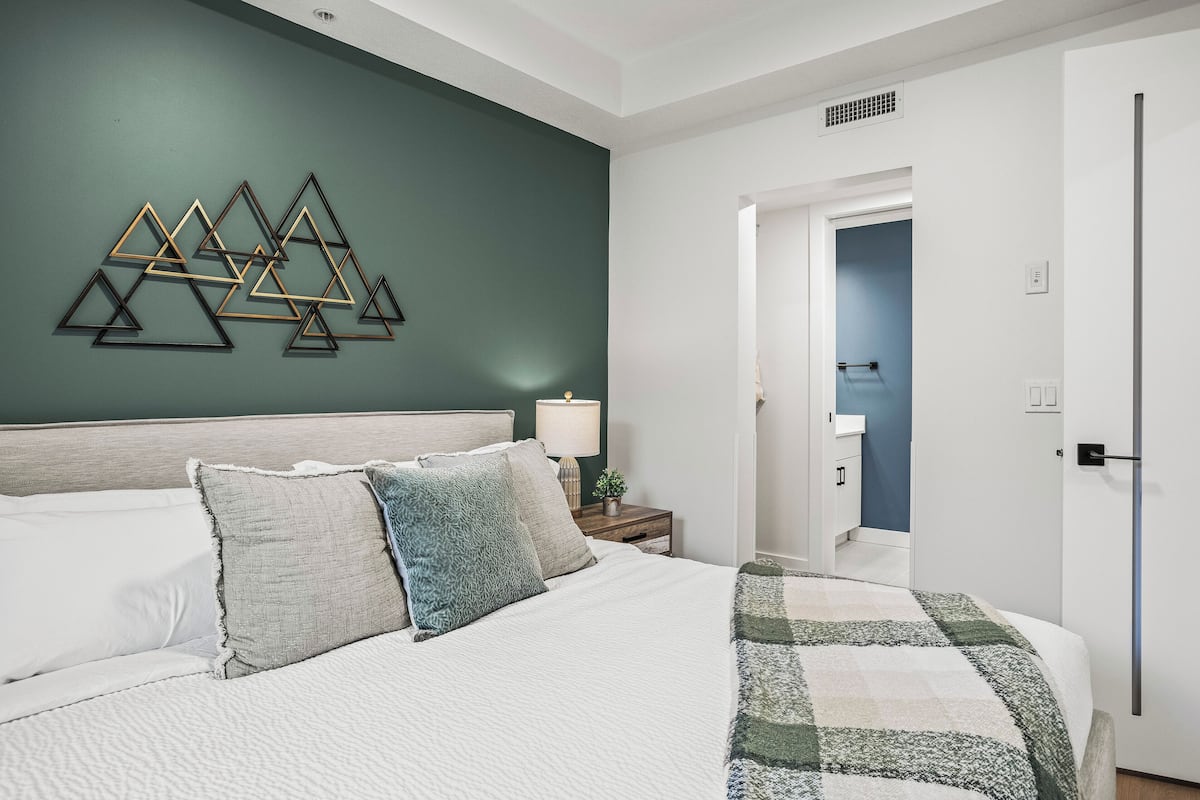 A bedroom with a bed, pillows, a wall art piece, a nightstand with a lamp, and a door leading to another room.