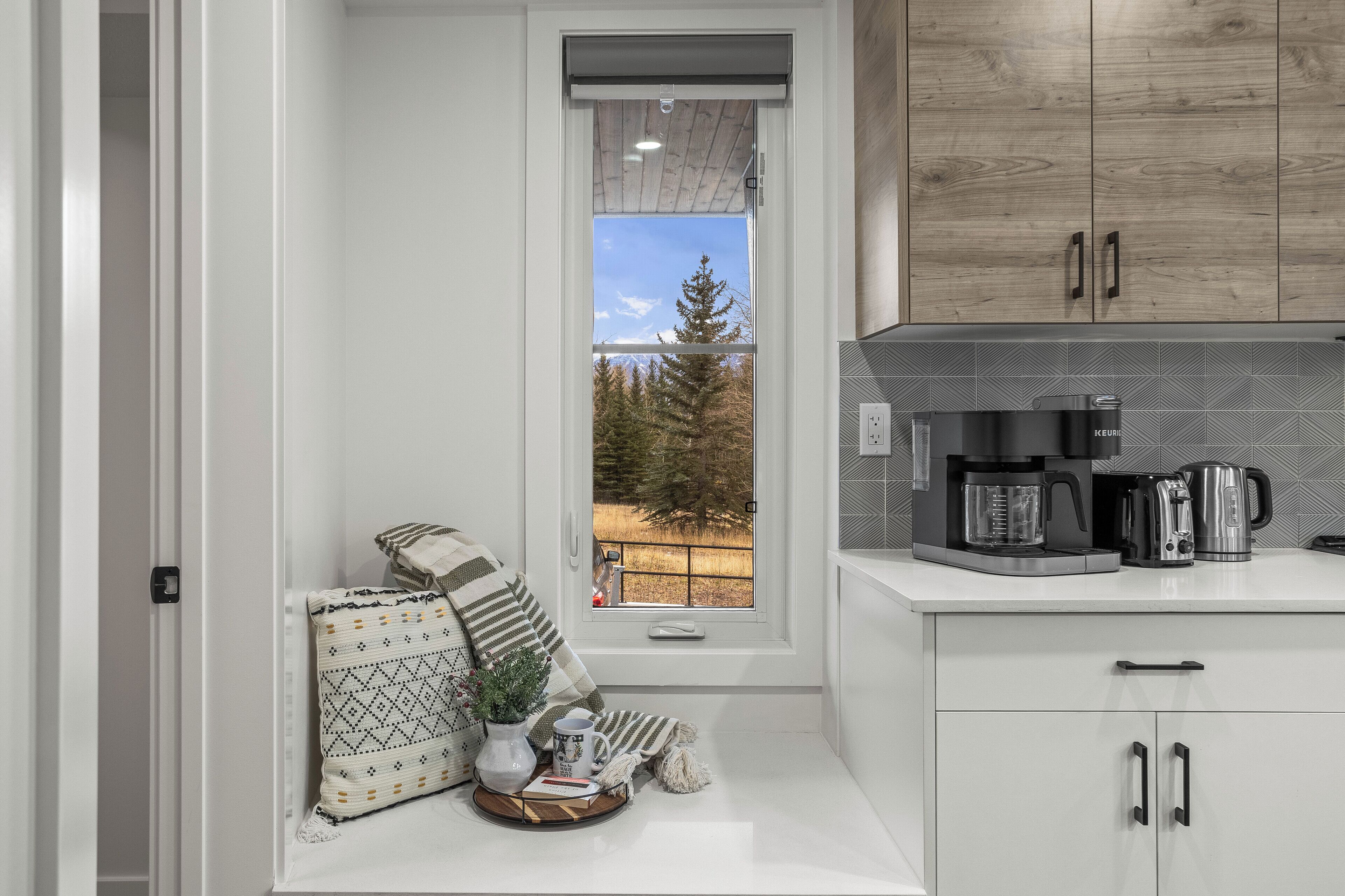 A modern kitchen with white cabinetry, a coffee maker, and a view of a forest through a window.