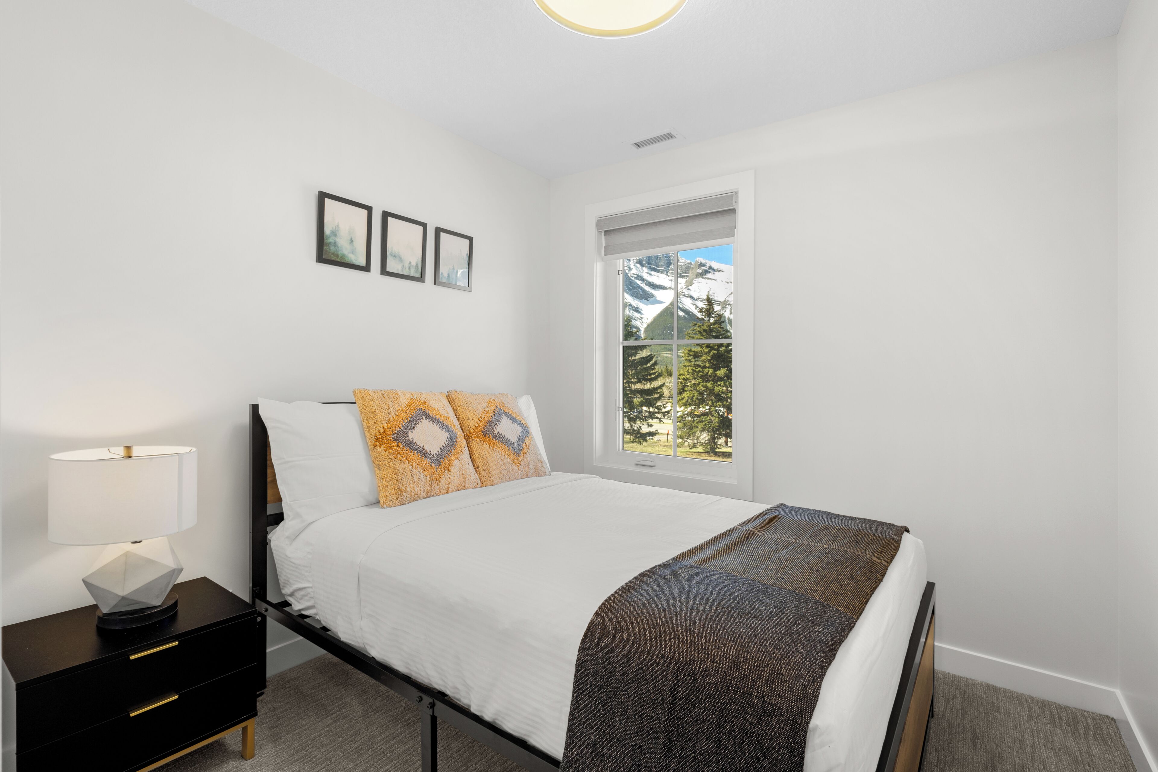 A bedroom with a bed, a nightstand, a window with a view of mountains, and framed pictures on the wall.