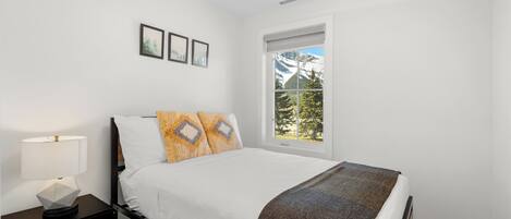 A bedroom with a bed, a nightstand, a window with a view of mountains, and framed pictures on the wall.