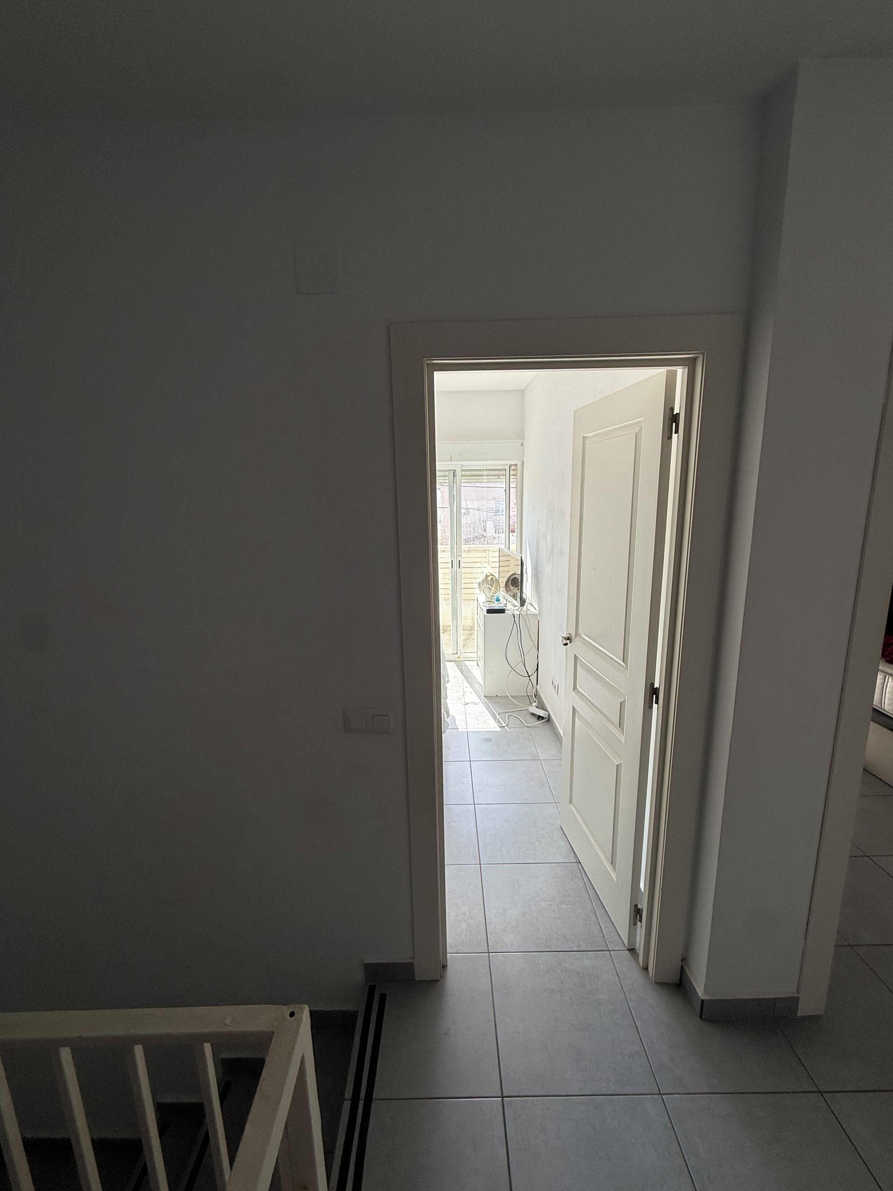Een gang met een witte deur die leidt naar een kamer met een raam en een betegelde vloer.
