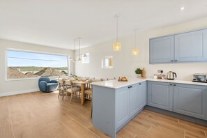 A modern kitchen with a large island, light blue cabinets, pendant lighting, and a dining area with a view of the countryside. - New for 2026  Rosemerryn , Holiday Home Padstow with breathtaking estuary views (Padstow)