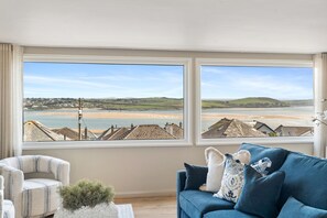 A room with a large window overlooking a beach and hills, a blue sofa, and a striped armchair. - New for 2026  Rosemerryn , Holiday Home Padstow with breathtaking estuary views (Padstow)