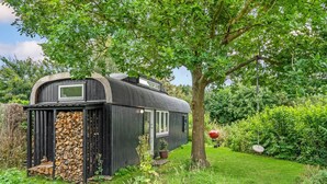 A black RV-style building with a window, a stack of firewood, and a barbecue grill in a lush garden. - 1 bedroom gorgeous home in Gilleleje (Gilleleje)