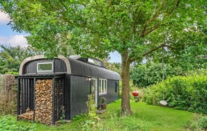 Ein schwarzes, mobilheimähnliches Gebäude mit einem Fenster, einem Holzstapel und einem Grill in einem üppigen Garten.