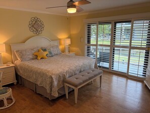Ein Schlafzimmer mit einem Bett, Nachttischlampen, einer Bank, einem Deckenventilator und Blick durch große Fenster.
