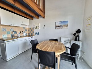 A compact kitchen with white cabinets, a wooden table, black chairs, a coffee machine, and a painting of a beach. - Charming apartment ideal for 4 people, perfect for a comfortable stay between ocean and... (BISCARROSSE PLAGE)