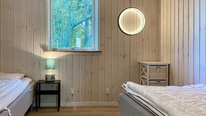 Et soveværelse med to senge, et vindue med udsigt til træer, et sengebord med en lampe og et lille skab.