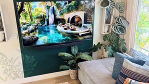 Ein Wohnzimmer mit einem Fernseher, der eine Naturdarstellung zeigt, einer Couch, einem Couchtisch mit einem Buch und Zimmerpflanzen.