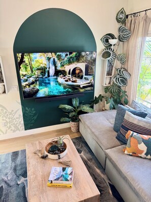 Ein Wohnzimmer mit einem Fernseher, der eine Naturdarstellung zeigt, einer Couch, einem Couchtisch mit einem Buch und Zimmerpflanzen.